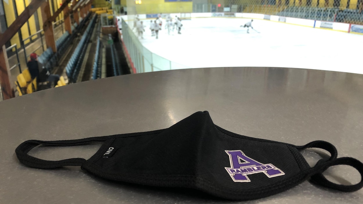Un masque, avec le logo de l'équipe, est sur une table. À l'arrière-plan, les joueurs sont sur la patinoire.