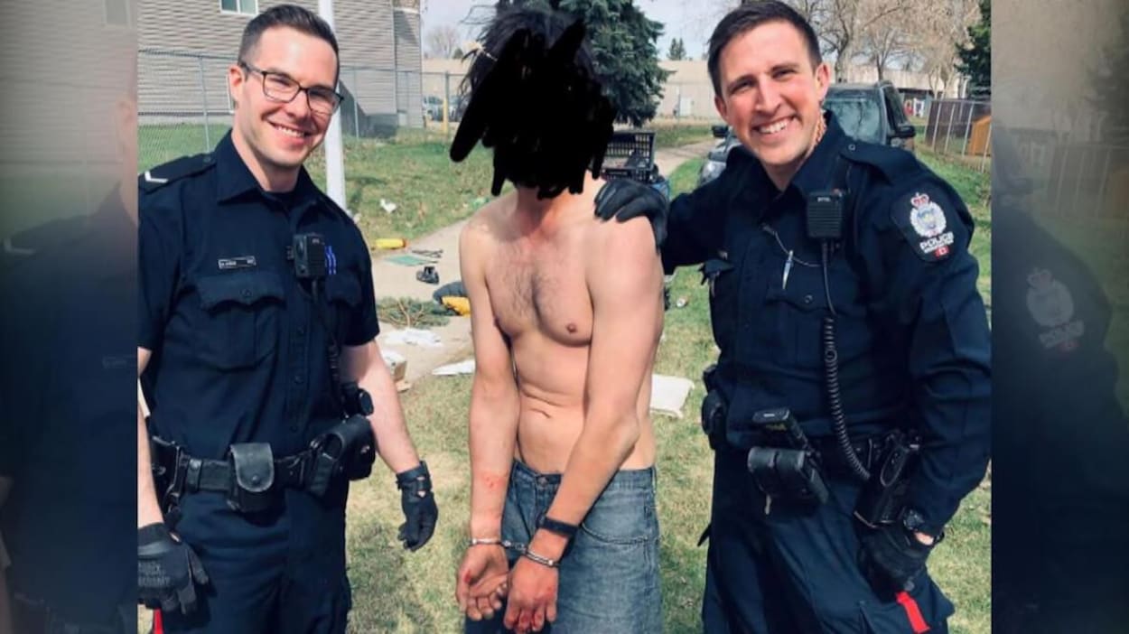 Deux policiers en uniforme sourient autour d'un homme menotté et torse nu. Le visage de l'homme menotté est noirci pour cacher son identité.