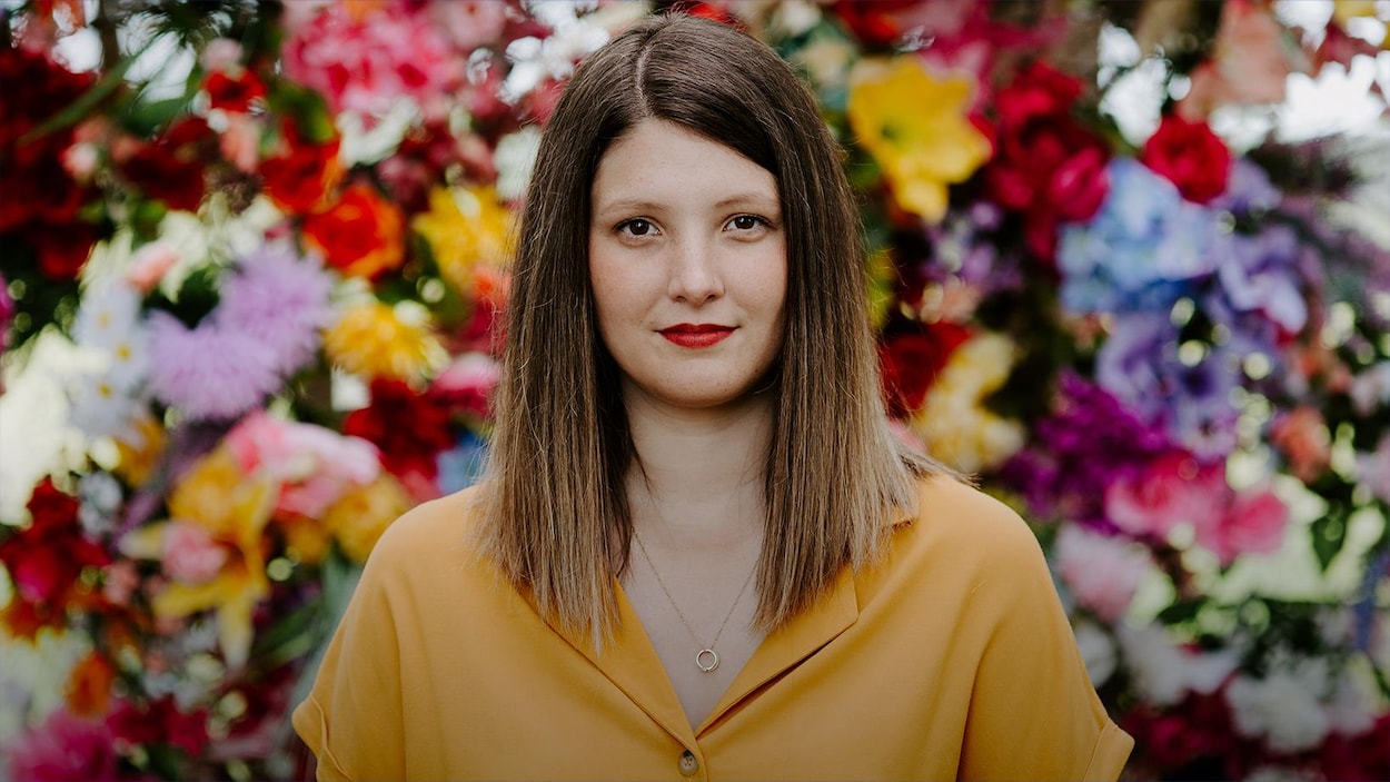 Chloé Breault pour "Plage des morons", son premier album complet (2020) : elle fixe l'objectif; cheveux droits; chemise jaune; elle se tient devant un mur de fleurs colorées.