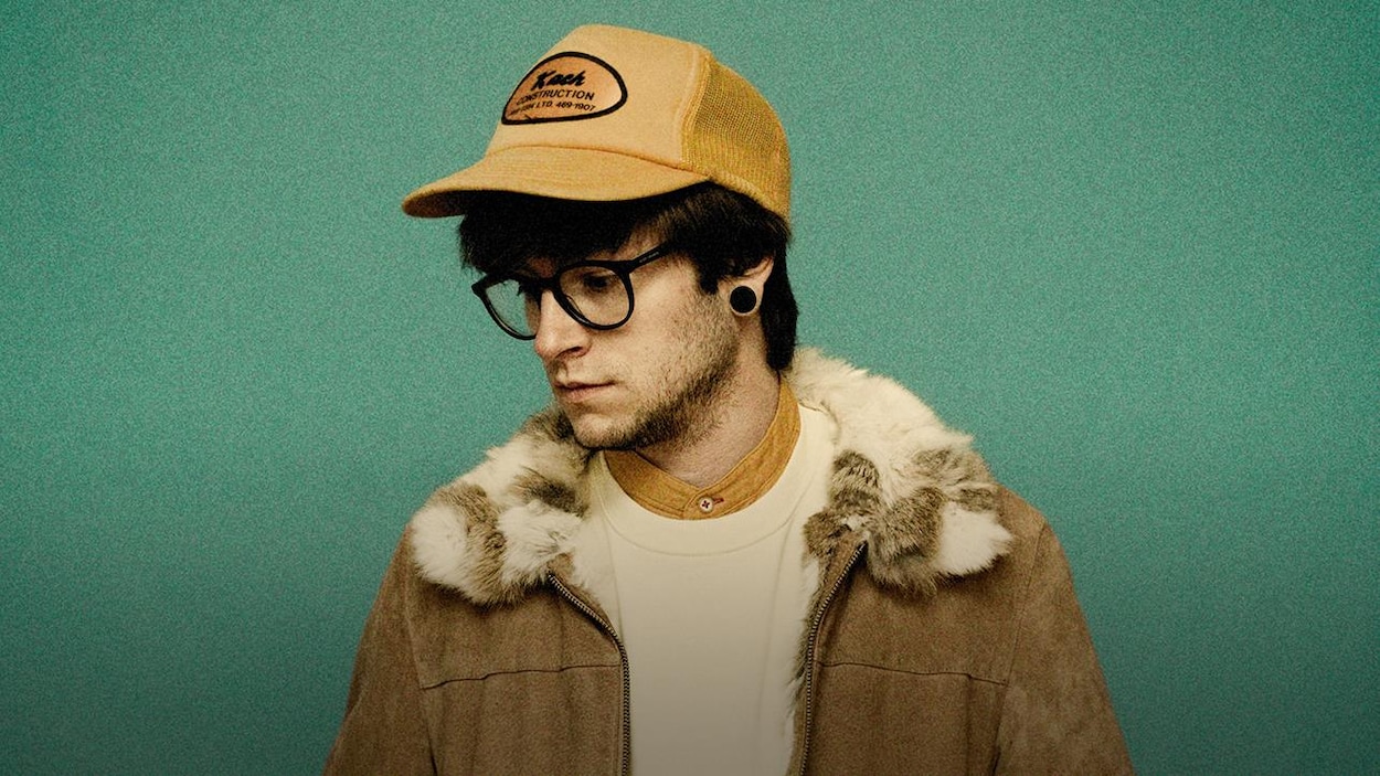 Pierre Guitard portant des lunettes et une casquette devant un mur vert