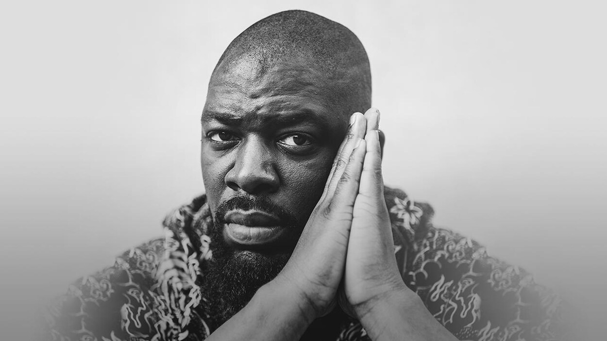 Le rappeur Dramatik pose sans sourire, les deux mains jointes.
