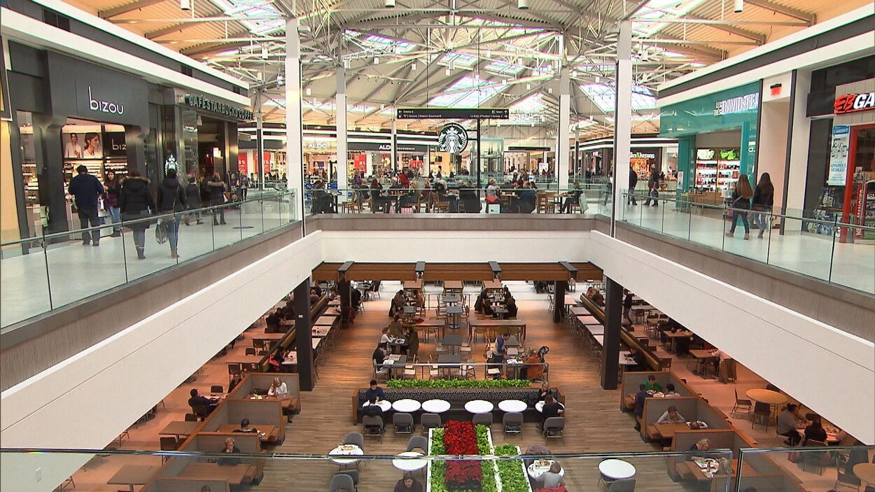Des gens circulent dans un centre commercial à deux étages. En bas, on voit une série de tables et des arrangements floraux, tandis qu'en haut, un café Starbucks trône au centre des différentes boutiques.