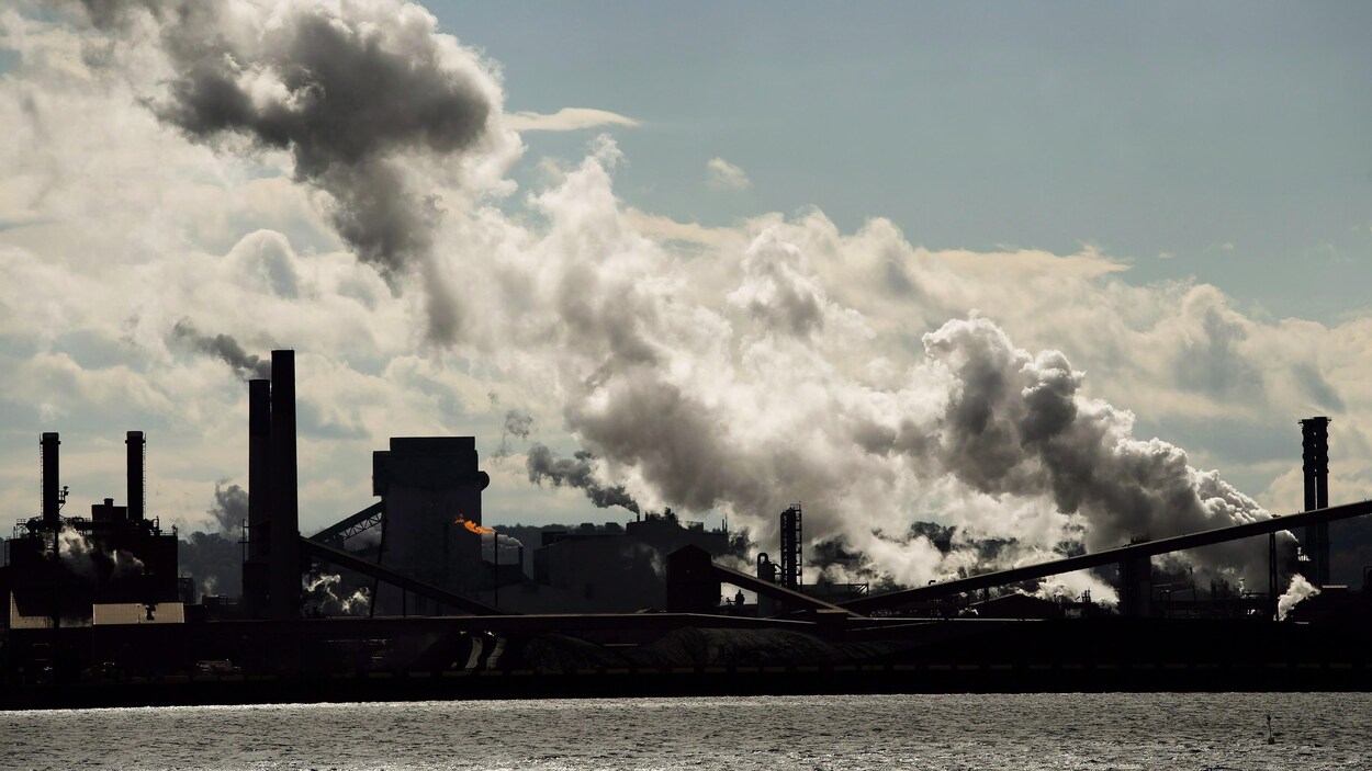 Une usine sur le bord de l'eau. De grandes volutes de fumée s'échappent et assombrissent le ciel.