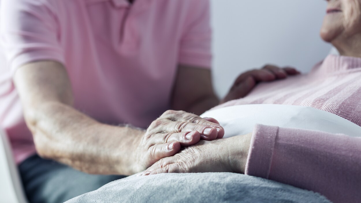 Une personne âgée est couchée et un homme lui tient la main.
