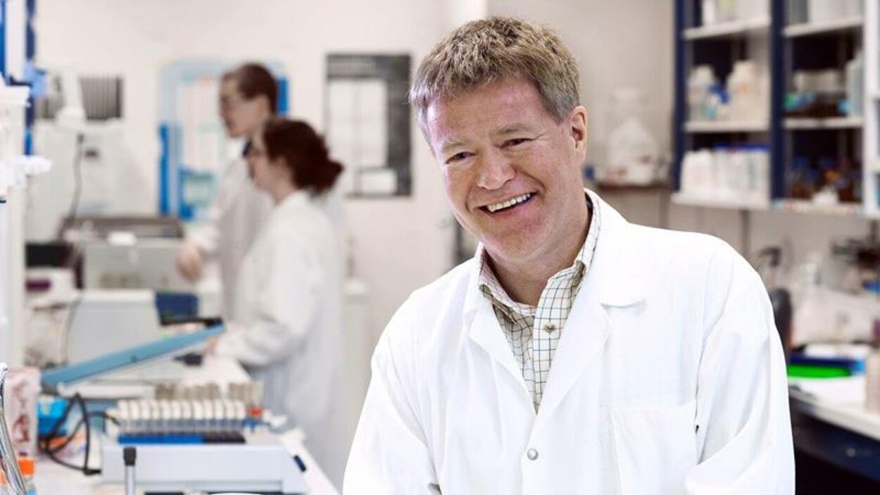 Un homme souriant et portant un sarrau blanc se trouve dans un laboratoire de recherche scientifique.