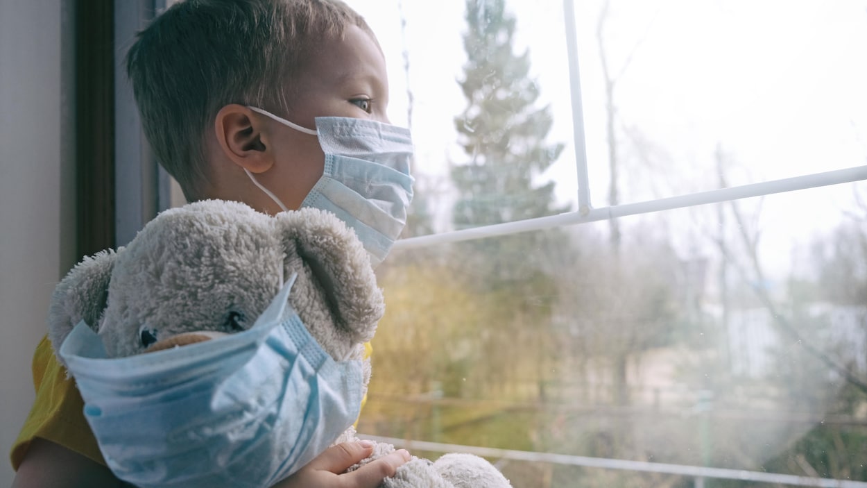 Un enfant avec un masque regarde par la fenêtre avec son toutou.