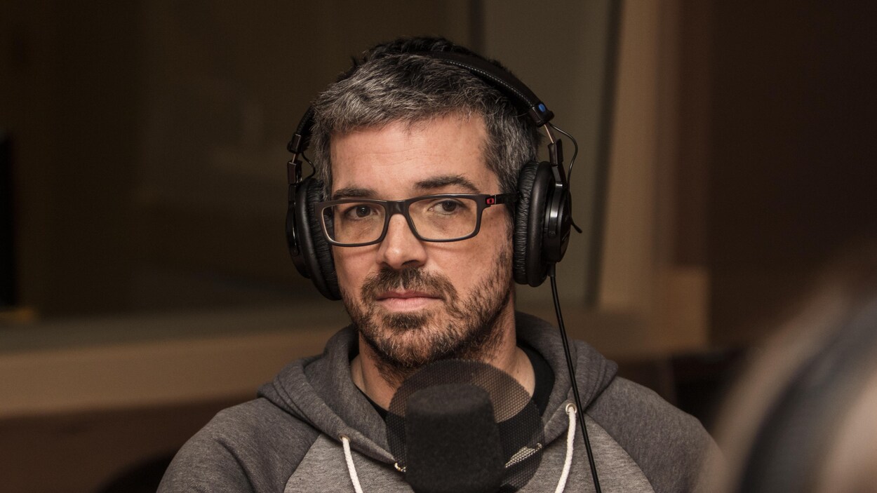 L'homme a les cheveux et la barbe poivre et sel et porte des lunettes rectangulaires