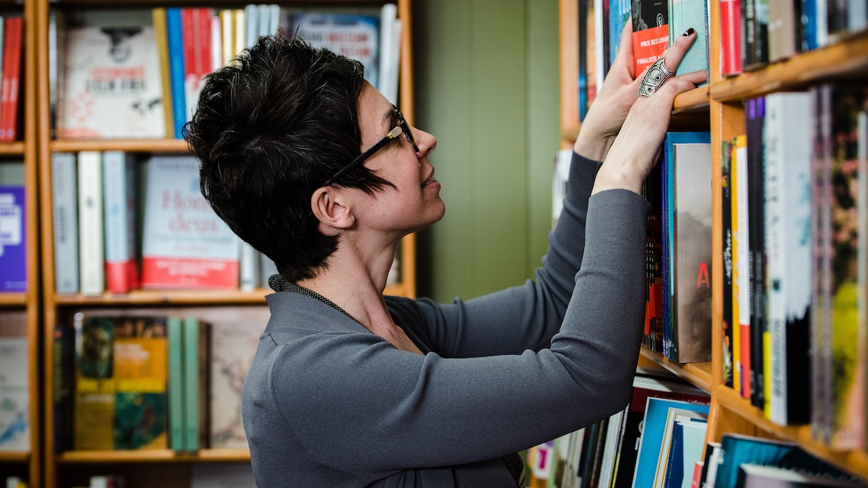 On voit une femme qui se trouve dans une bibliothèque. Elle manipule quelques livres dans une étagère.