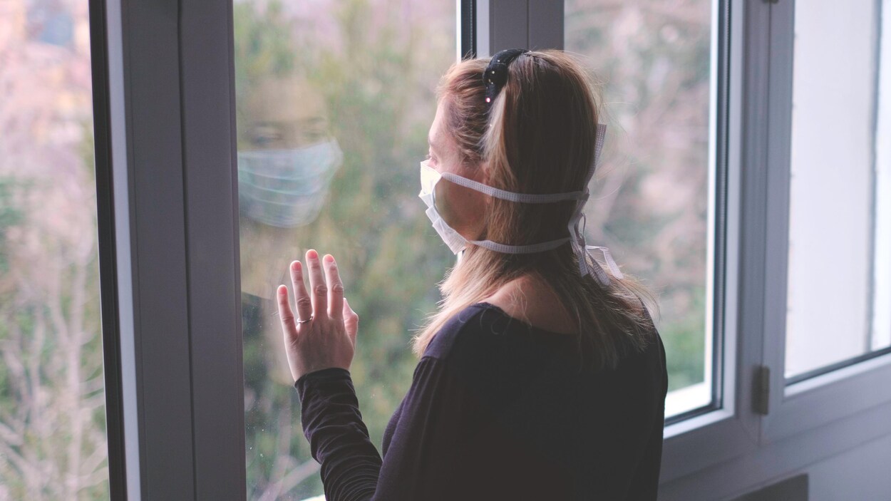 Une femme portant un masque en train de regarder par la fenêtre.