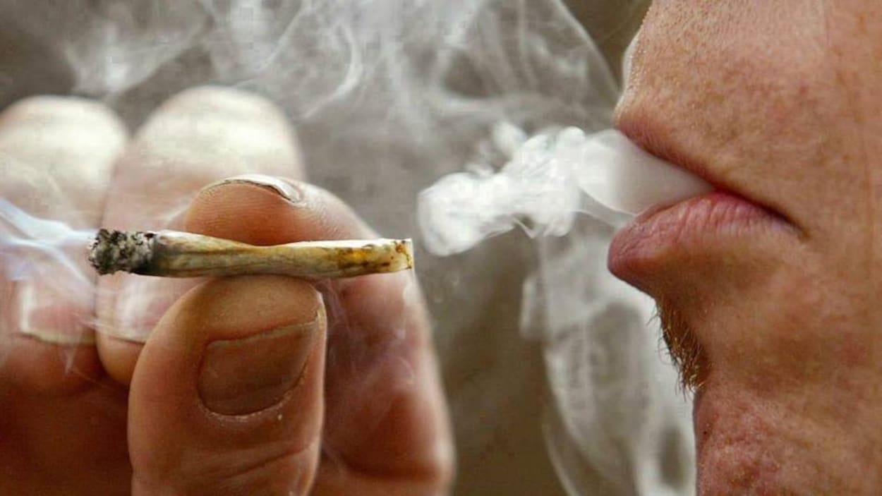 Un homme tient un joint, et vient de prendre une bouffée de marijuana, et de la fumée sort de sa bouche.