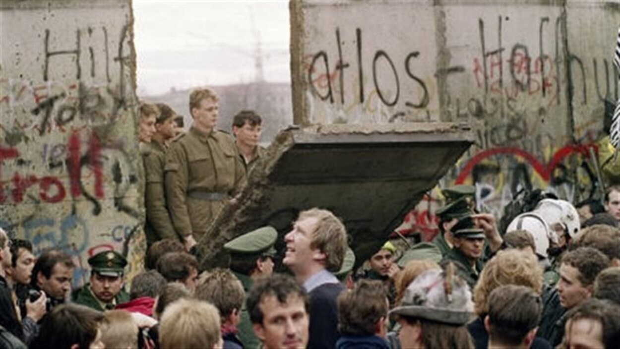 Des soldats sont debout sur un des panneaux tombés du mur de Berlin et regardent la foule massée tout près.