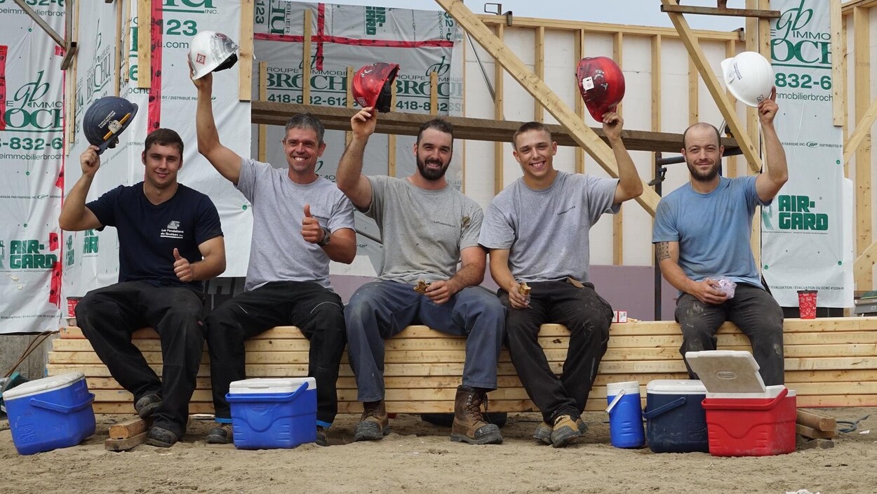 Cinq travailleurs de la construction lèvent leur chapeau en souriant.