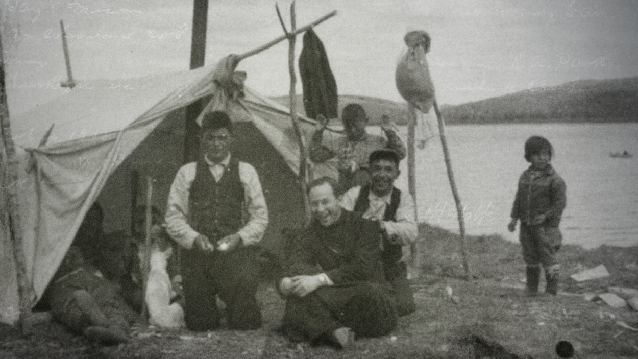 Le père Alexis Joveneau avec une famille innue. Ils sont tous assis près d'une tente, au bord de l'eau.