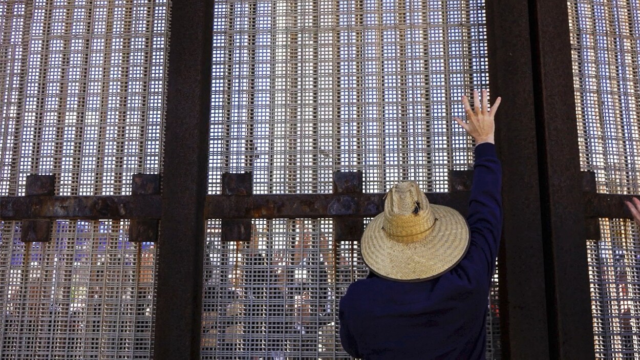 Une personne avec un chapeau de paille se tient debout devant une partie du mur entre le Mexique et les États-Unis