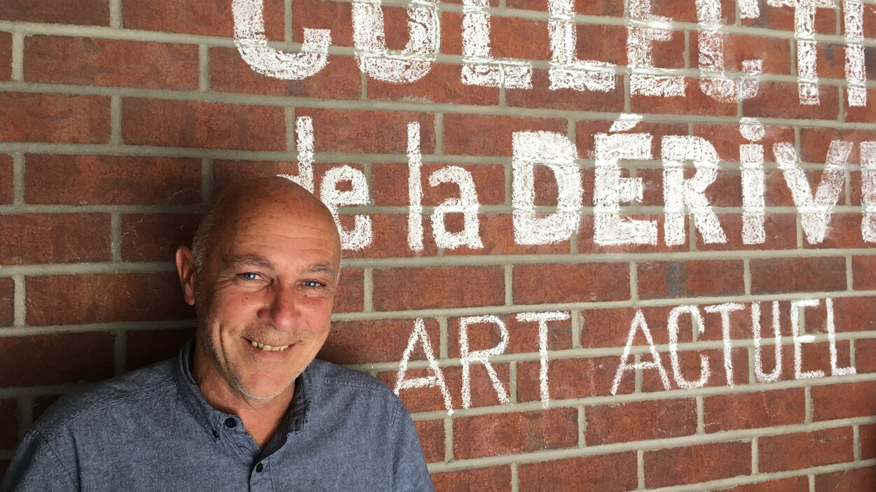 Richard Ferron souriant, adossé à un mur de briques où des mots sont peints en blanc.