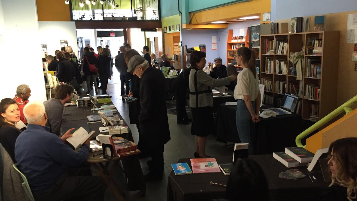 Une salle avec des tables et des personnes qui sont assises. On peut voir des bibliothèques et des livres.