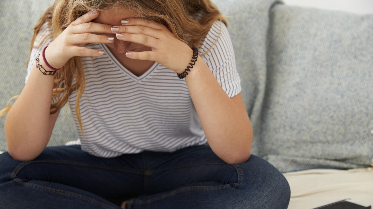 Une jeune fille est assise sur un sofa et elle se tient la tête dans les mains.