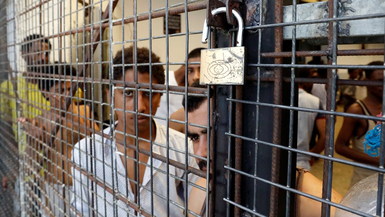 Des migrants emprisonnés regardent le photographe derrière des barreaux et une porte maintenue fermée par un cadenas. Des migrants emprisonnés regardent le photographe derrière des barreaux et une porte maintenue fermée par un cadenas.