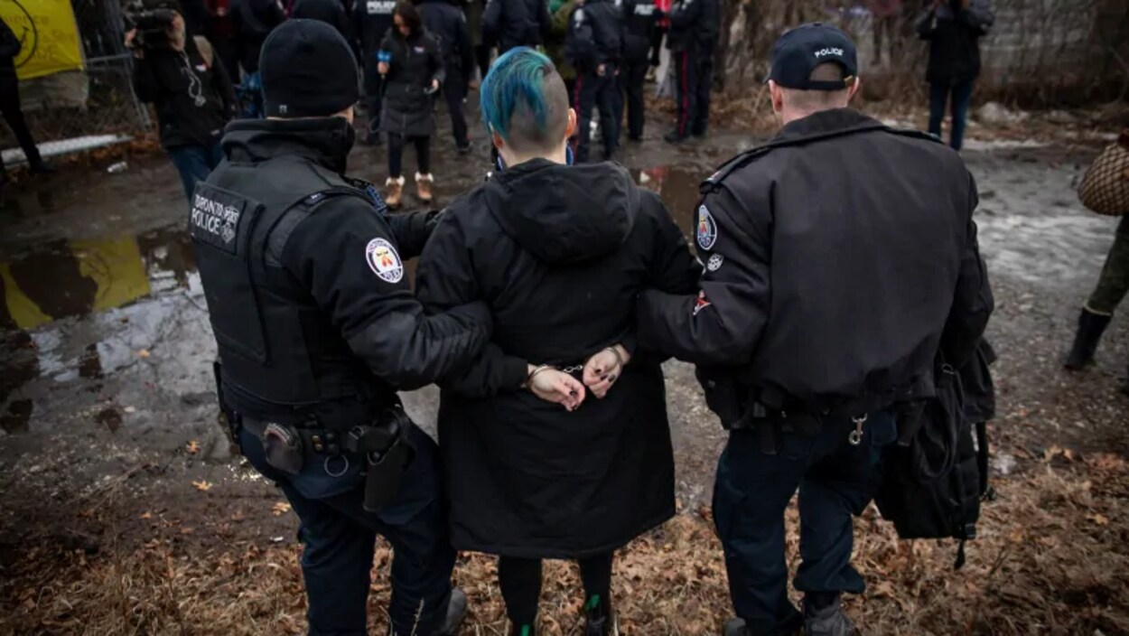 Une femme menottée est tenue par les bras par deux officiers de police.