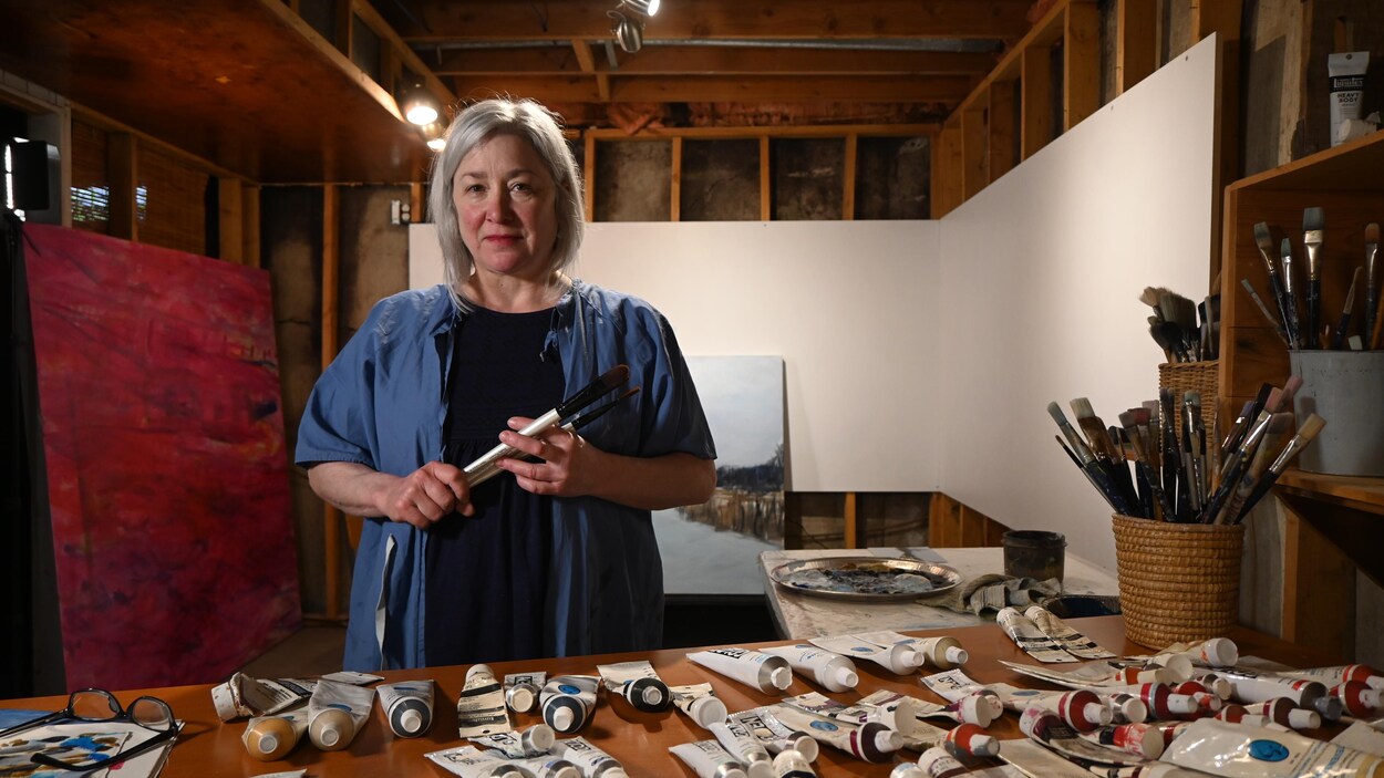 Brigitte Dion, pinceaux à la main, est debout devant ses tubes de peinture acrylique
