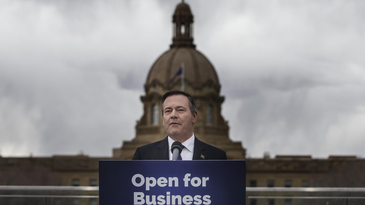 Jason Kenney se tient au podium devant un panneau "open for Business". Il est devant l'Assemblée législative de l'Alberta.