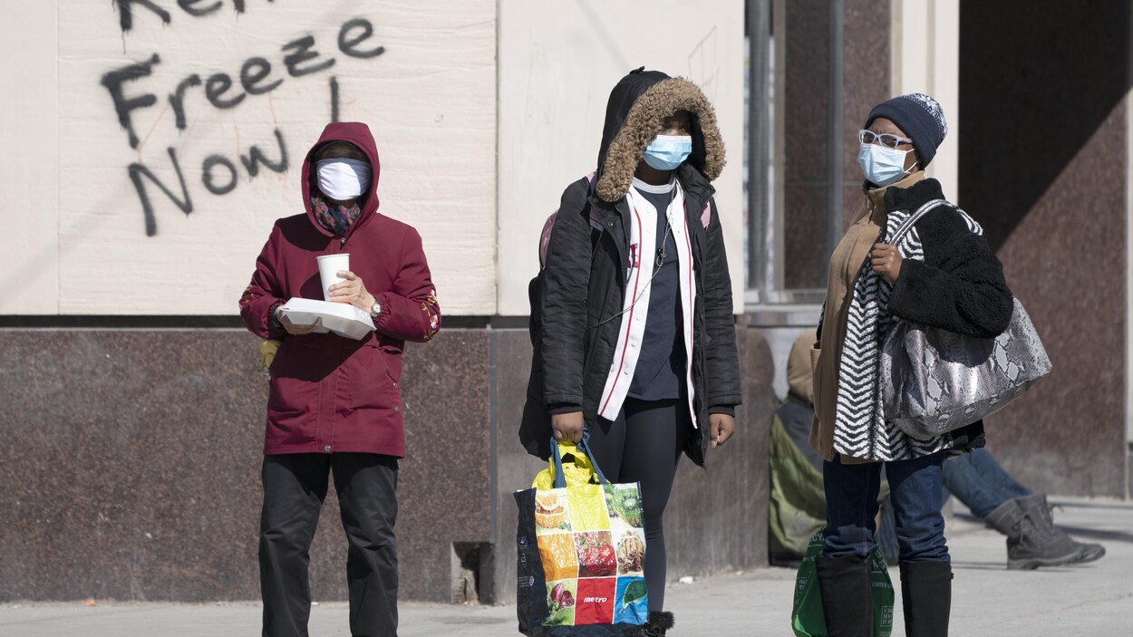 Des piétons masqués patientent sur un trottoir devant un graffiti demandant un gel des loyers.