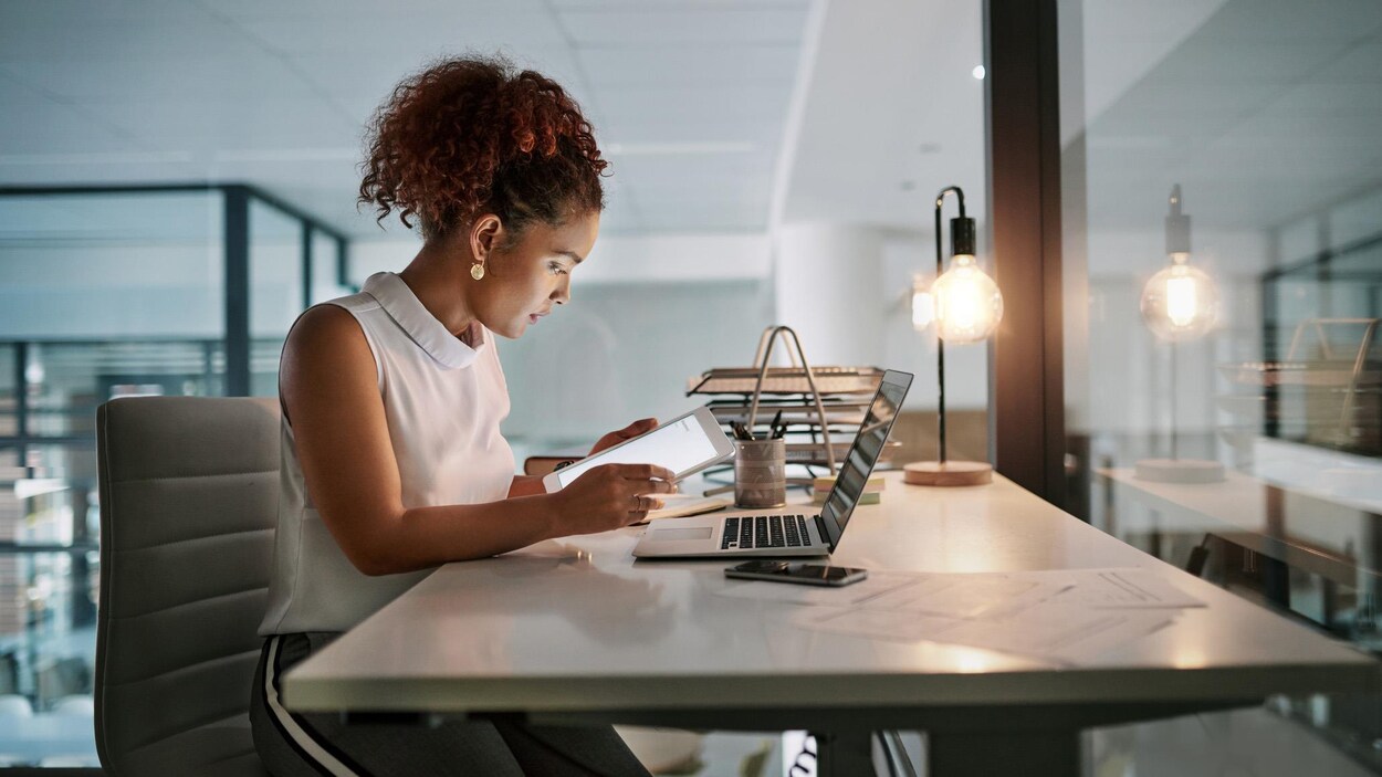 Une femme travaille sur une tablette informatique; un ordinateur est posé sur son bureau.