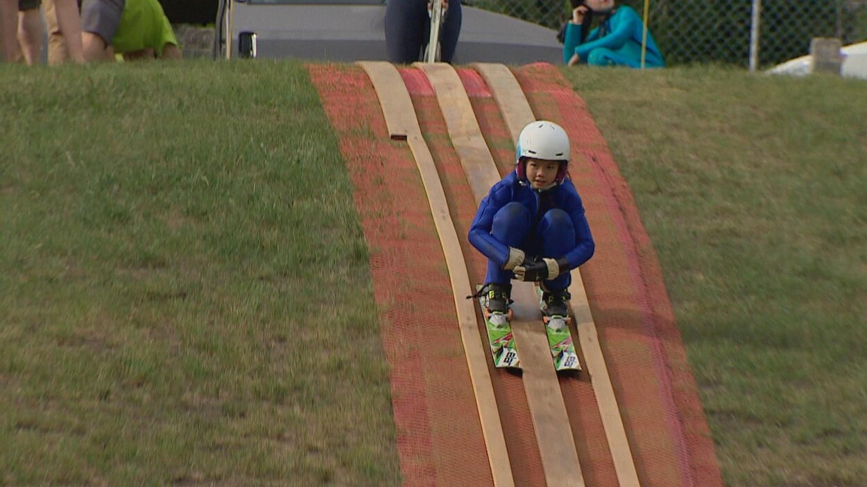Une enfant vêtue d'une combinaison de ski, d'un casque et de gants est accroupie sur ses skis alors qu'elle descend une piste installée sur de l'herbe.