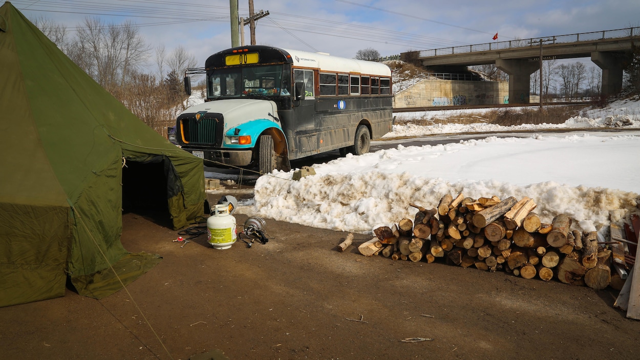 Un camion de transport de personnes fait face à une tente verte. On remarque des bûches de bois et une bouteille de gaz. Le tout forme un campement, au second plan, un pont supportant une voie ferrée du CN.