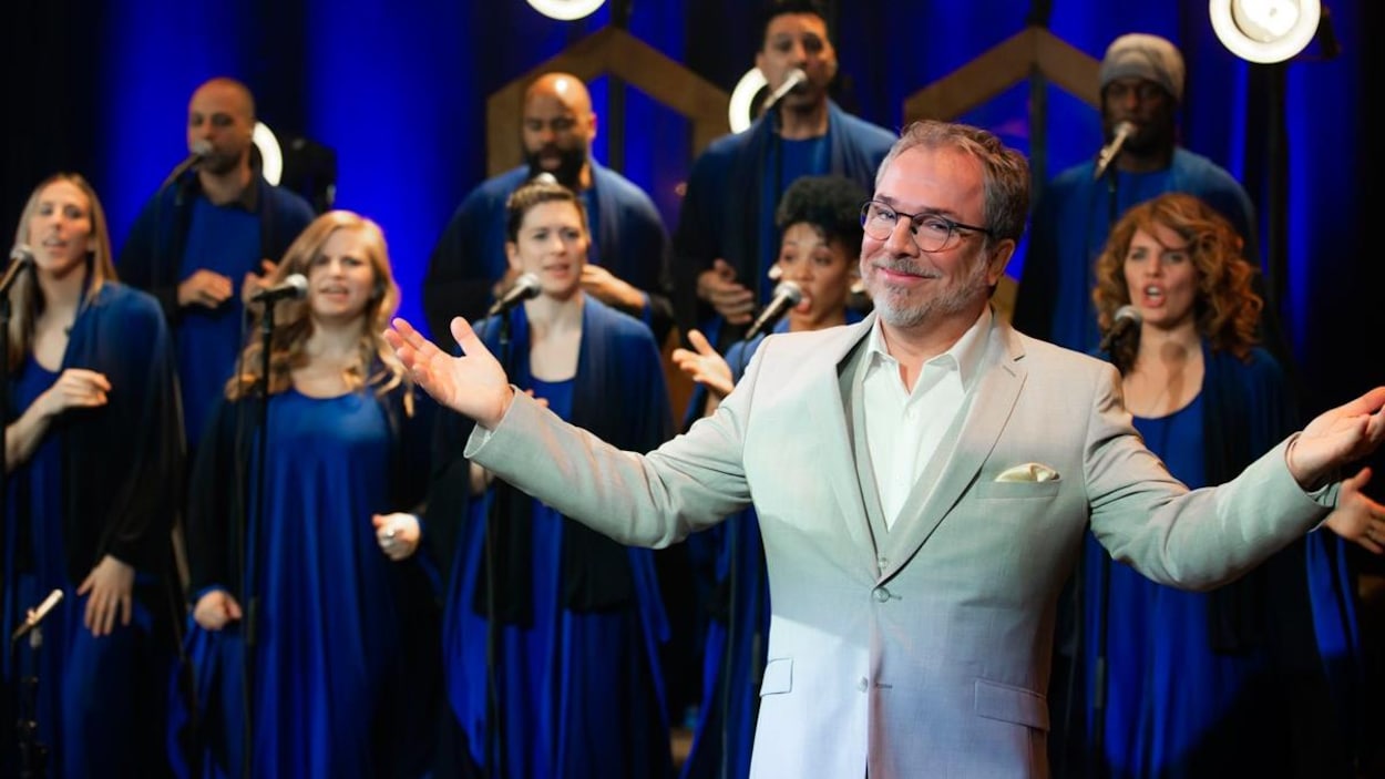 Christian Bégin pose les bras ouverts devant la chorale, dont les membres sont habillés en bleu.