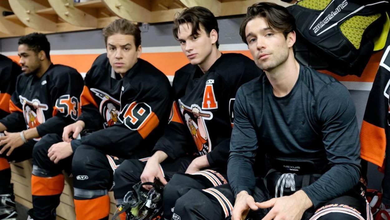 Des comédiens vêtus avec de l'équipement de hockey, sur le banc, dans une chambre des joueurs.