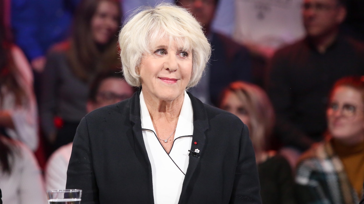 Ses cheveux sont blonds. Elle porte une chemise blanche et une veste noire.