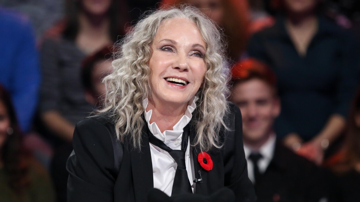 Elle porte une chemise blanche, une cravate noire et un veston noir sur un plateau de télévision. On voit un public derrière elle.