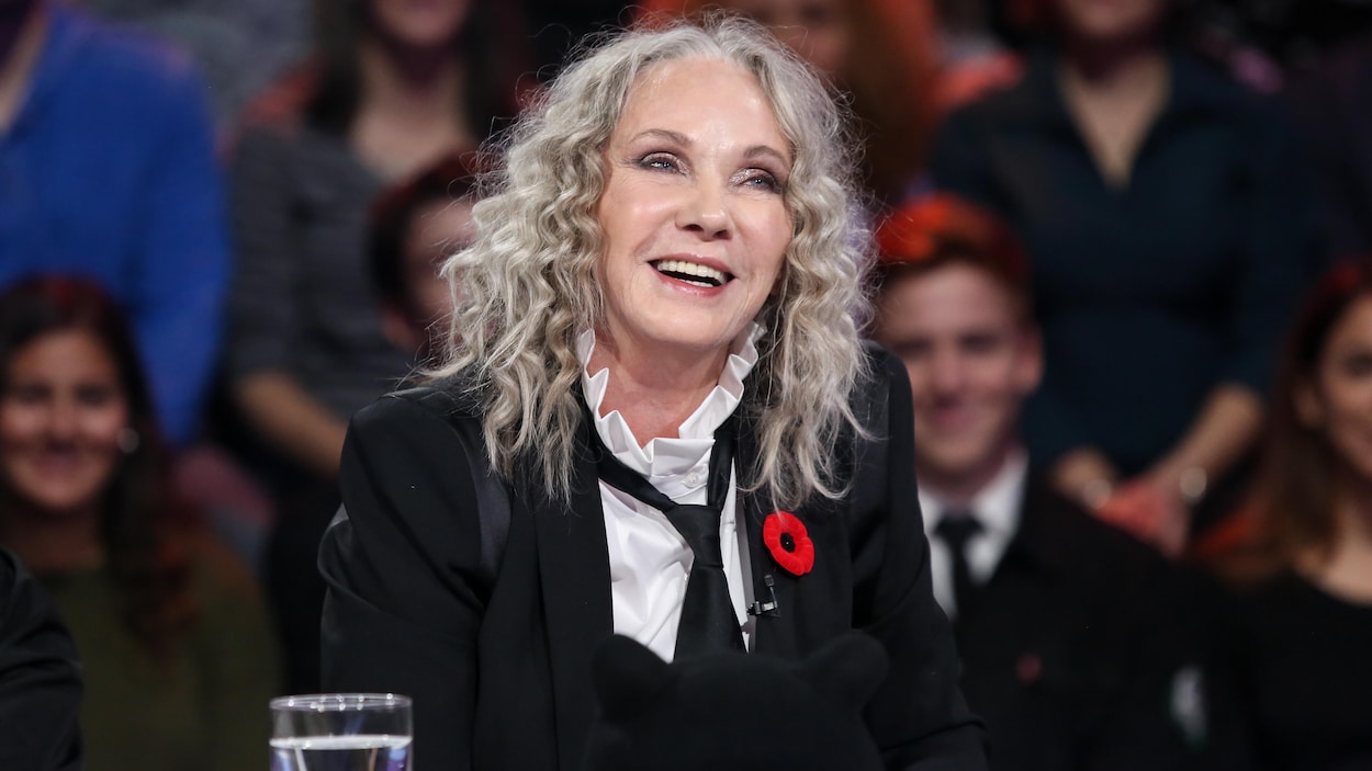 Elle porte une chemise blanche, une cravate noire et un veston noir.