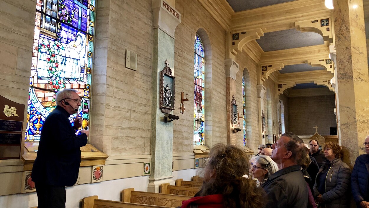 Un homme habillé en noir présente un vitrail de l'église. Des personnes l'écoutent attentivement. Un homme habillé en noir présente un vitrail de l'église. Des personnes l'écoutent attentivement.