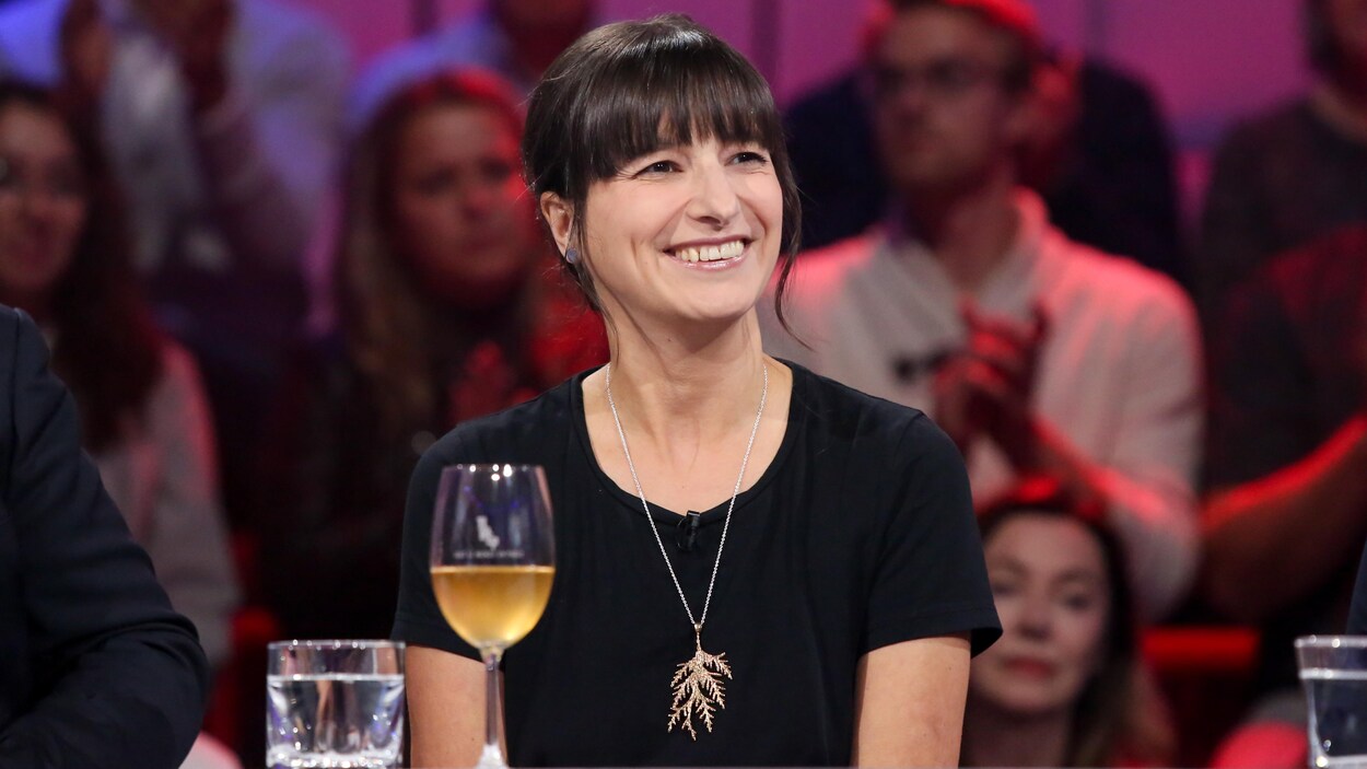 Elle sourit. Ses cheveux sont bruns. Elle porte un robe noire et un pendentif en argent.