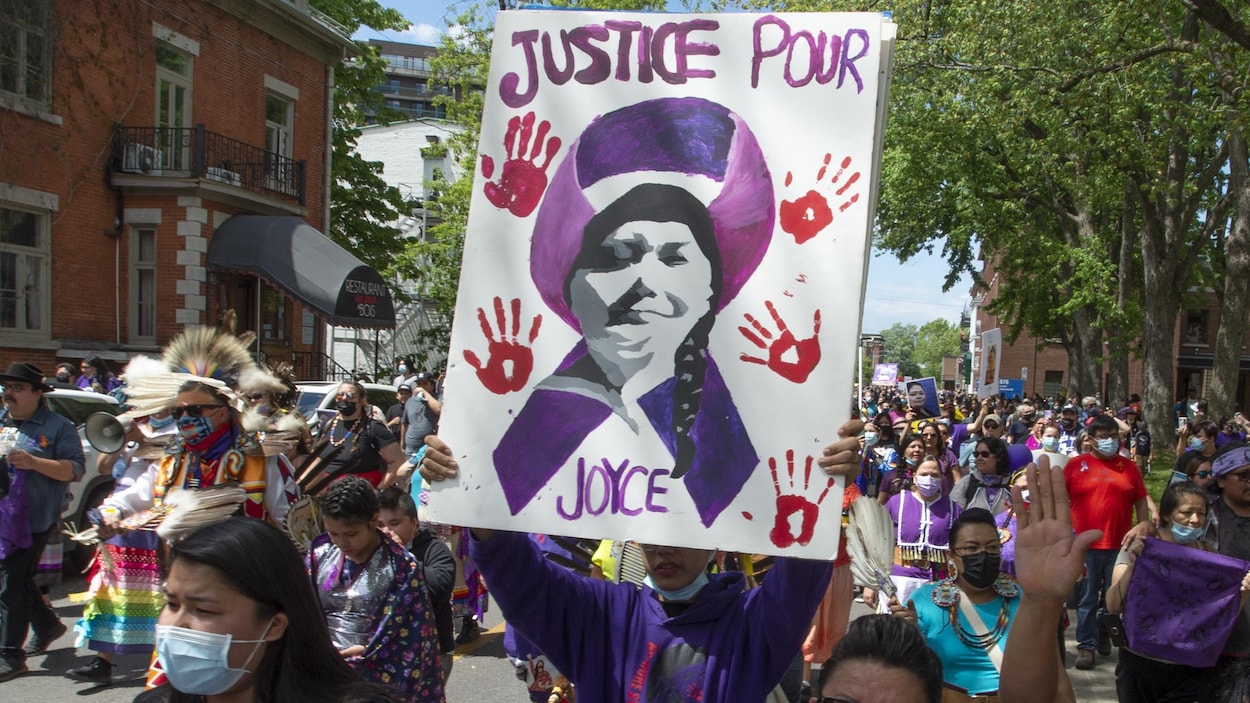 Des personnes manifestent dans la rue. Au centre de la photo, une pancarte est tenue par une personne. La pancarte est une peinture qui montre la face de Joyce Echaquan, superposée sur un ruban mauve. Les mots "Justice pour Joyce" sont également peints. Autour de la face de Joyce, des retrouve des traces de mains rouges, symbole de la lutte des femmes et filles autochtones disparues et assassinées.