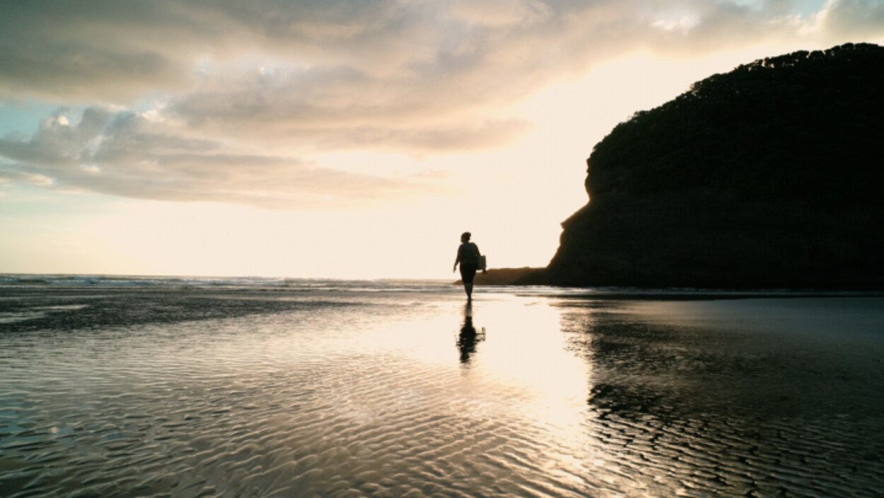 Une silhouette marche sur une plage, les pieds dans l'eau, alors que le soleil se couche.