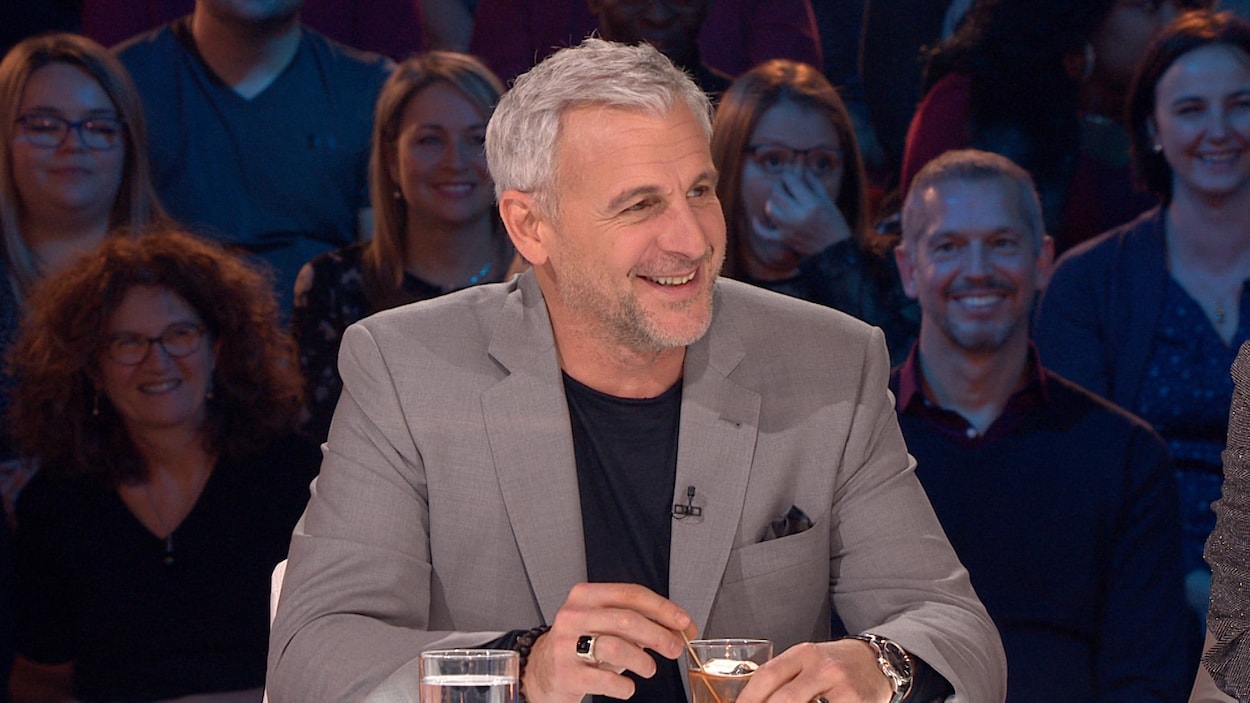 Patrick Huard porte une veste grise et un t-shirt noir.