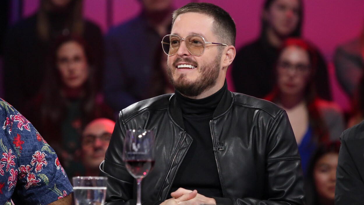 L'homme est assis à une table sur le plateau d'une émission de télévision, un verre de vin rouge devant lui. Il porte une veste de cuir noir et des lunettes.