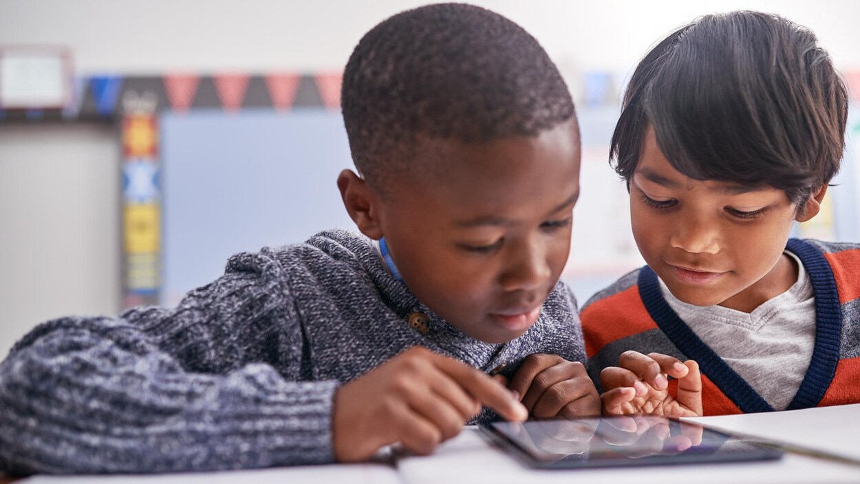 Deux jeunes garçons consultent une tablette dans une salle de cours. 