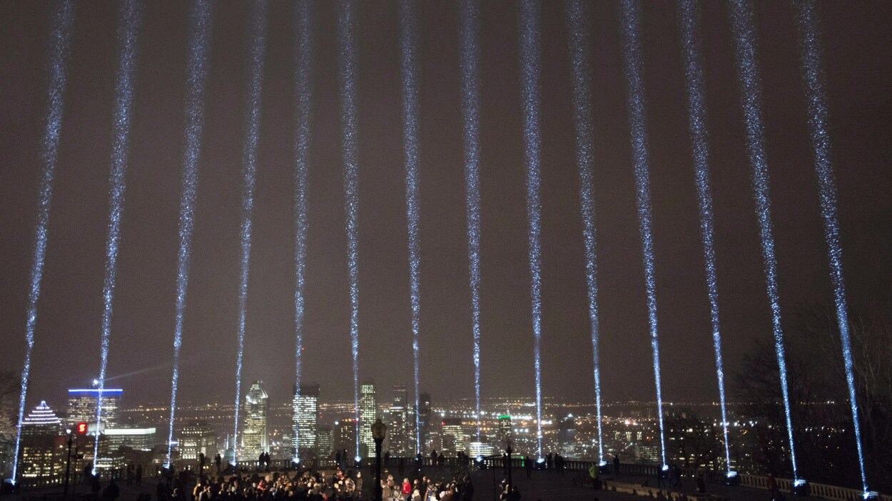 14 faisceaux lumineux qui pointent vers le ciel. Au loin, on peut voir la ville de Montréal la nuit. Dans le bas de l'image, des personnes sont rassemblées pour commémorer la tuerie à l'École polytechnique de Montréal.