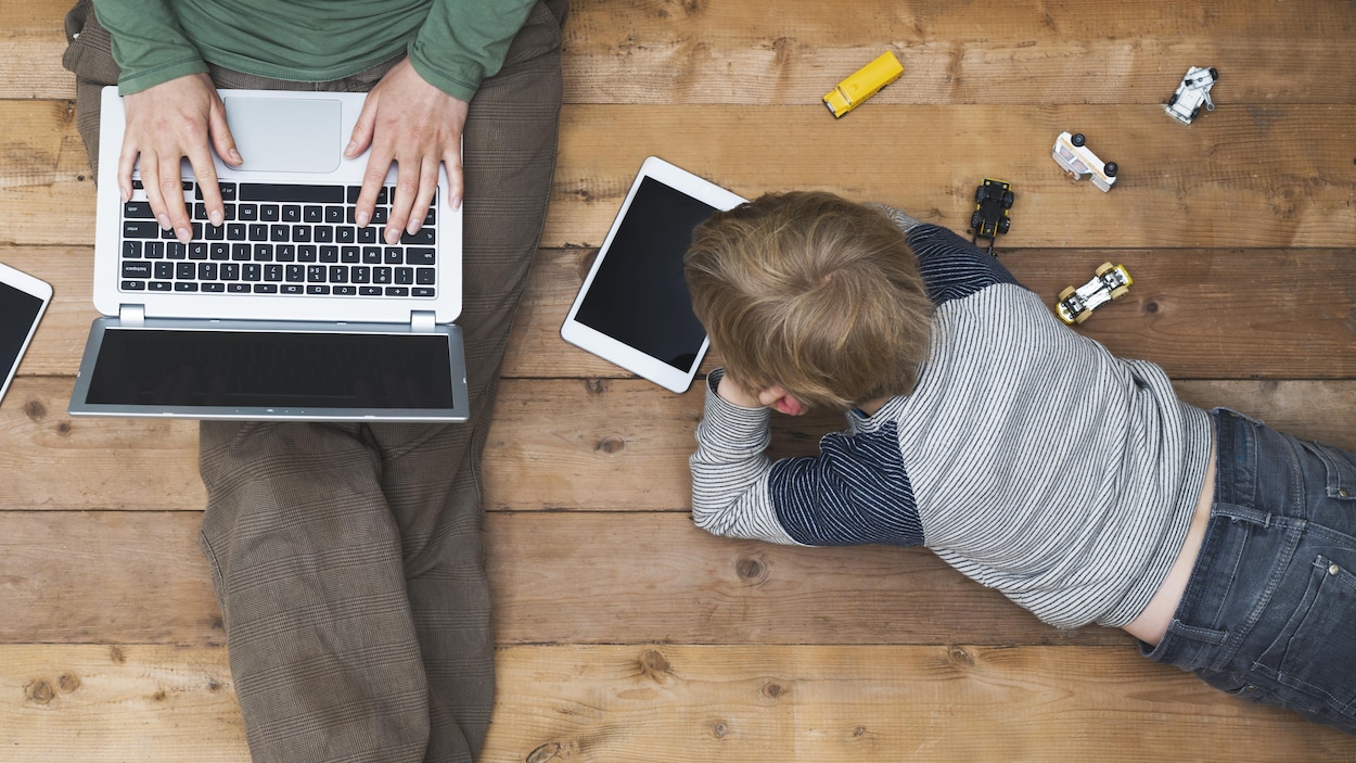 Une mère et son fils installés sur le plancher avec une collation, un ordinateur et une tablette.