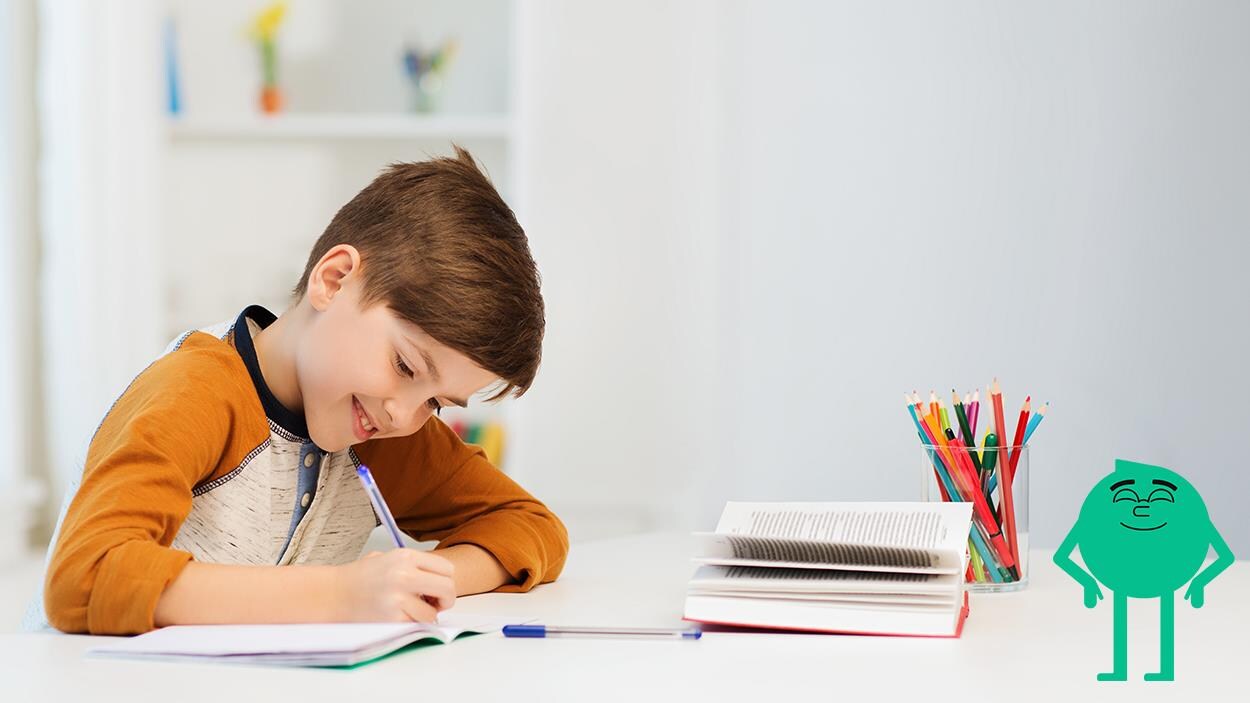 On voit une jeune garçon souriant attablé et écrivant dans son cahier scolaire