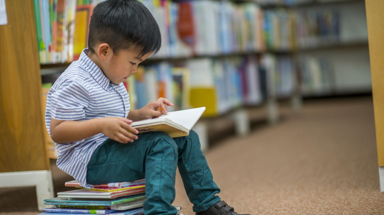 Un petit garçon asiatique assis sur une pile de livres, il lit dans la bibliothèque