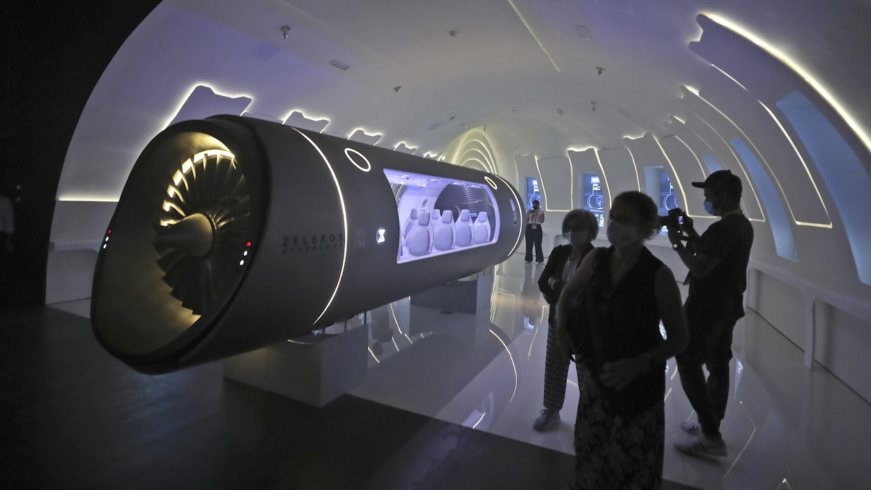 Trois personnes en silhouette observent un véhicule hyperloop dans une salle d'exposition.