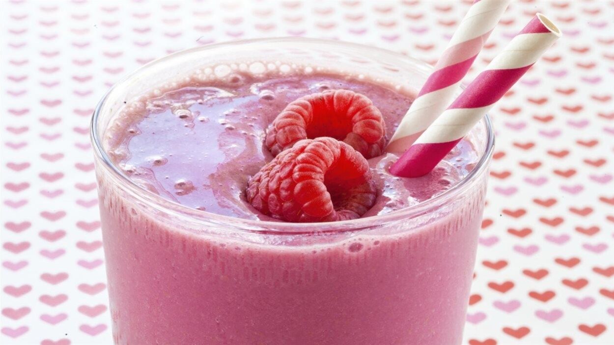 Gros plan sur un verre de smoothie orné de deux framboises et deux pailles sur une table dressée d'une nappe avec des coeurs.