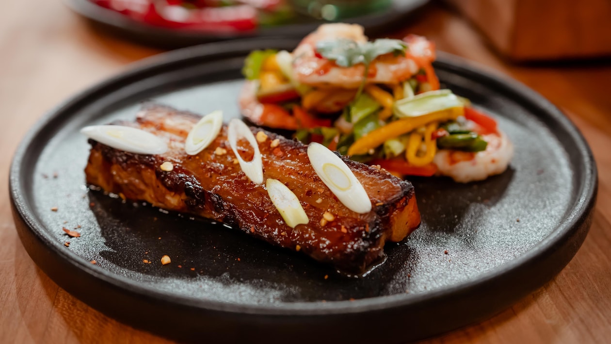 Un flanc de porc braisé accompagné d'une salade de crevettes dans une assiette noire.