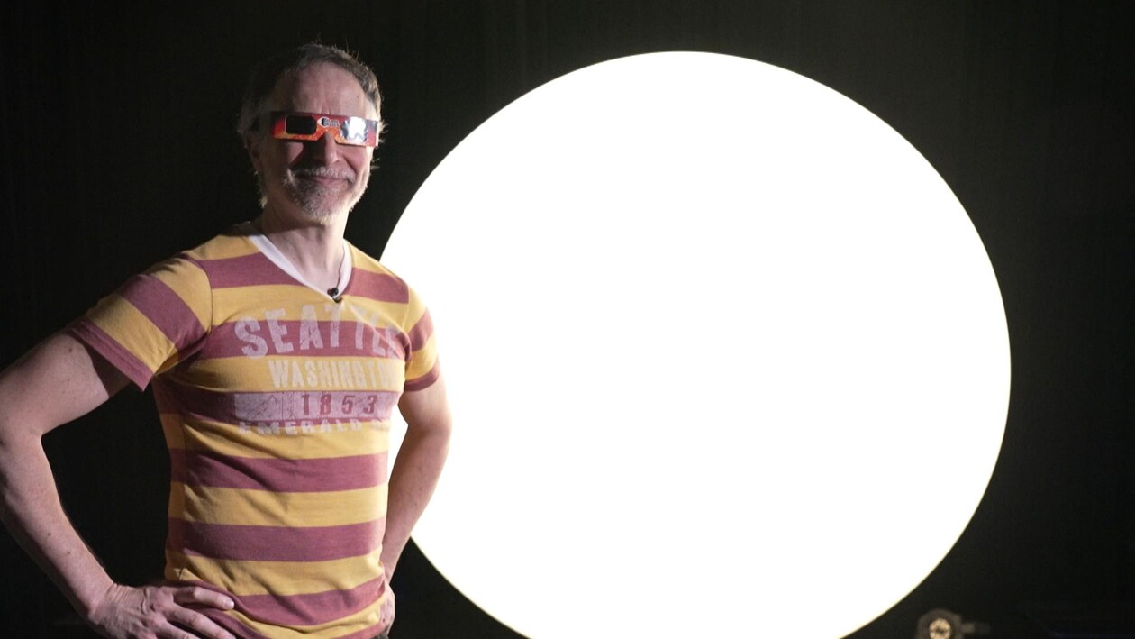Un homme sourit avec des lunettes conçues pour regarder une éclipse devant un grand ballon illuminé.
