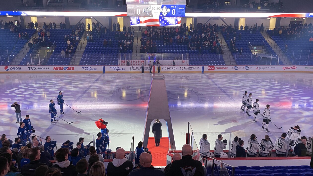 Des joueurs de hockey sur une patinoire et des spectateurs debout dans les gradins pendant qu'une femme est sur un tapis rouge sur la glace.